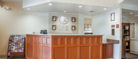 A hotel reception area with a wooden reception desk, a wall-mounted sign reading "Comfort Suites," and a bulletin board with various items.