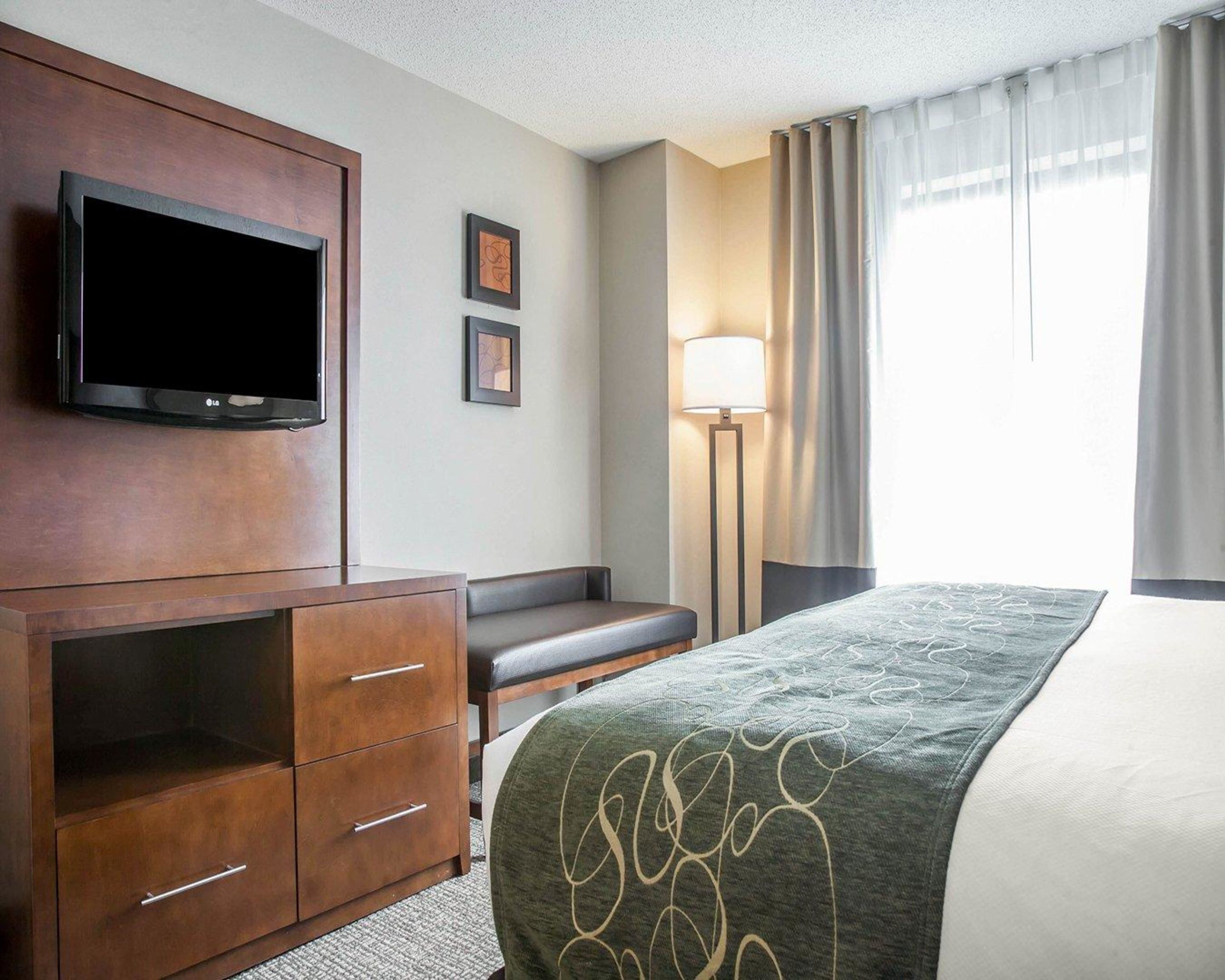 Une chambre d’hôtel équipée d’un téléviseur à écran plat fixé à un meuble en bois, d’un bureau avec une chaise, d’un lit recouvert d’une couverture verte, d’une lampe sur pied et d’une fenêtre avec des rideaux.