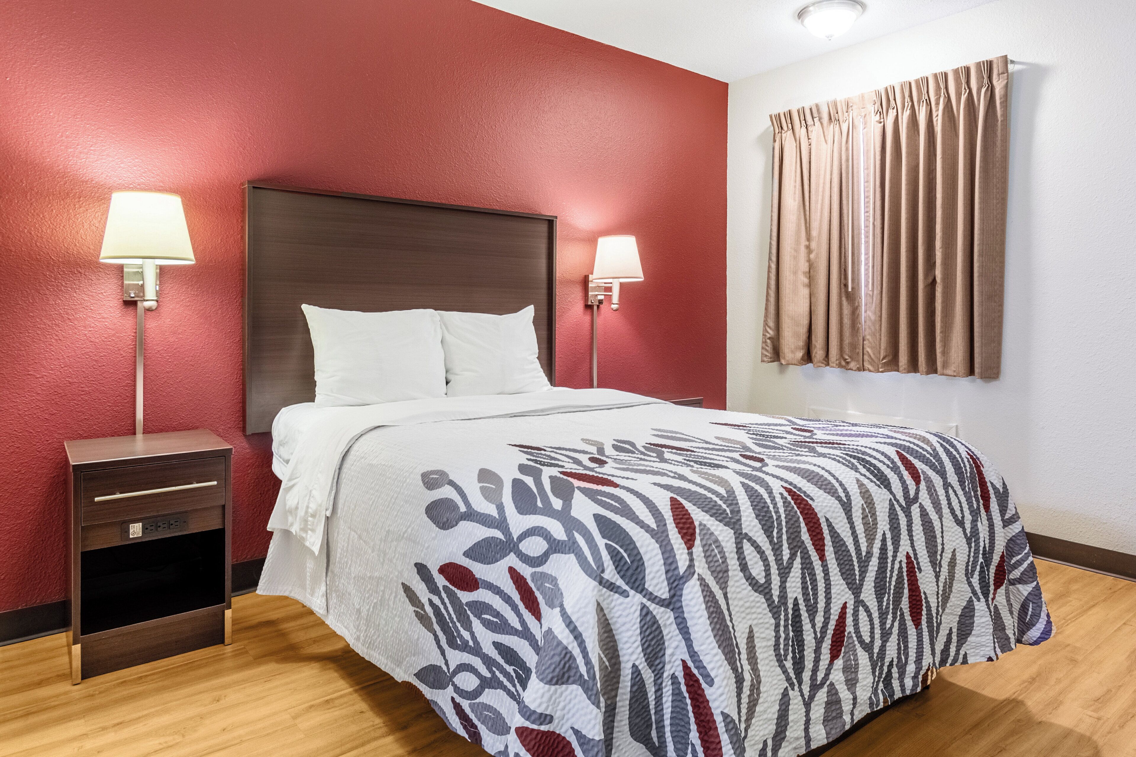 Habitación de hotel con cabecera de madera, dos lámparas de noche, una cama con colcha estampada, una mesita de noche y un ventanal con cortinas.