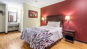 A hotel room with a bed, bedside lamps, a nightstand, a bathroom with a sink and mirror, and a framed picture on the wall. - Red Roof Inn Scottsburg (Scottsburg)