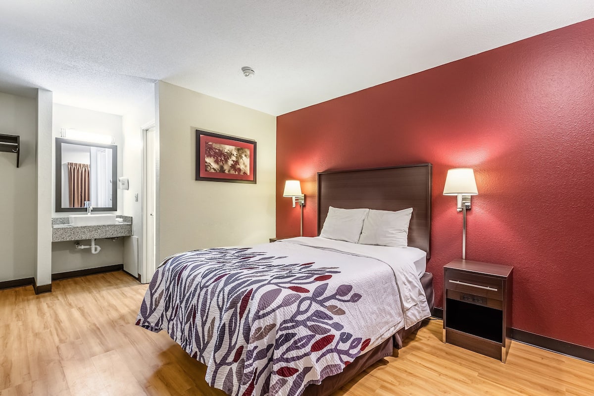 A hotel room with a bed, bedside lamps, a nightstand, a bathroom with a sink, and a framed picture on the wall.