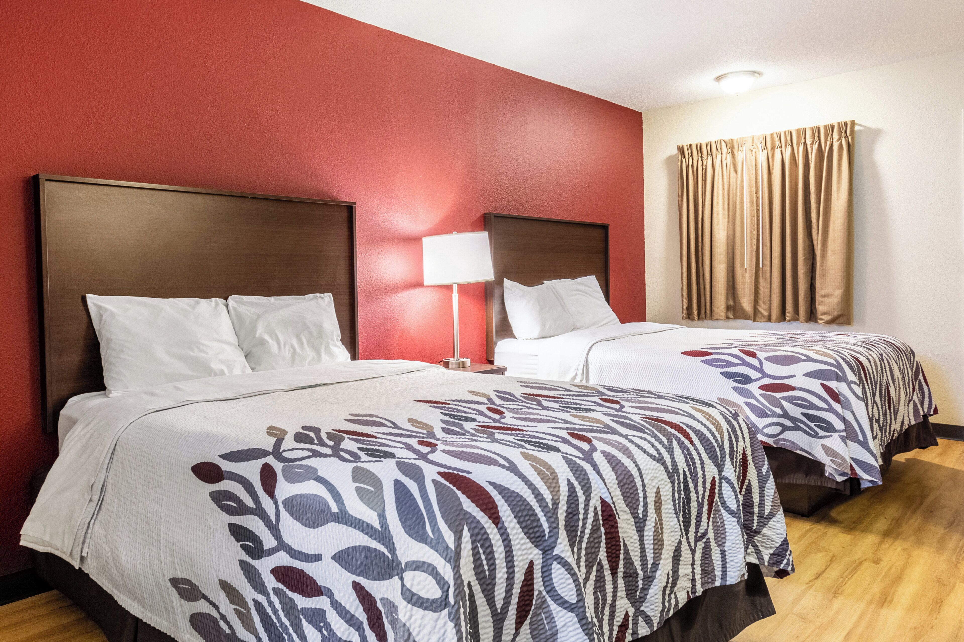 A hotel room with two beds, a red wall, a window with curtains, and a bedside lamp.