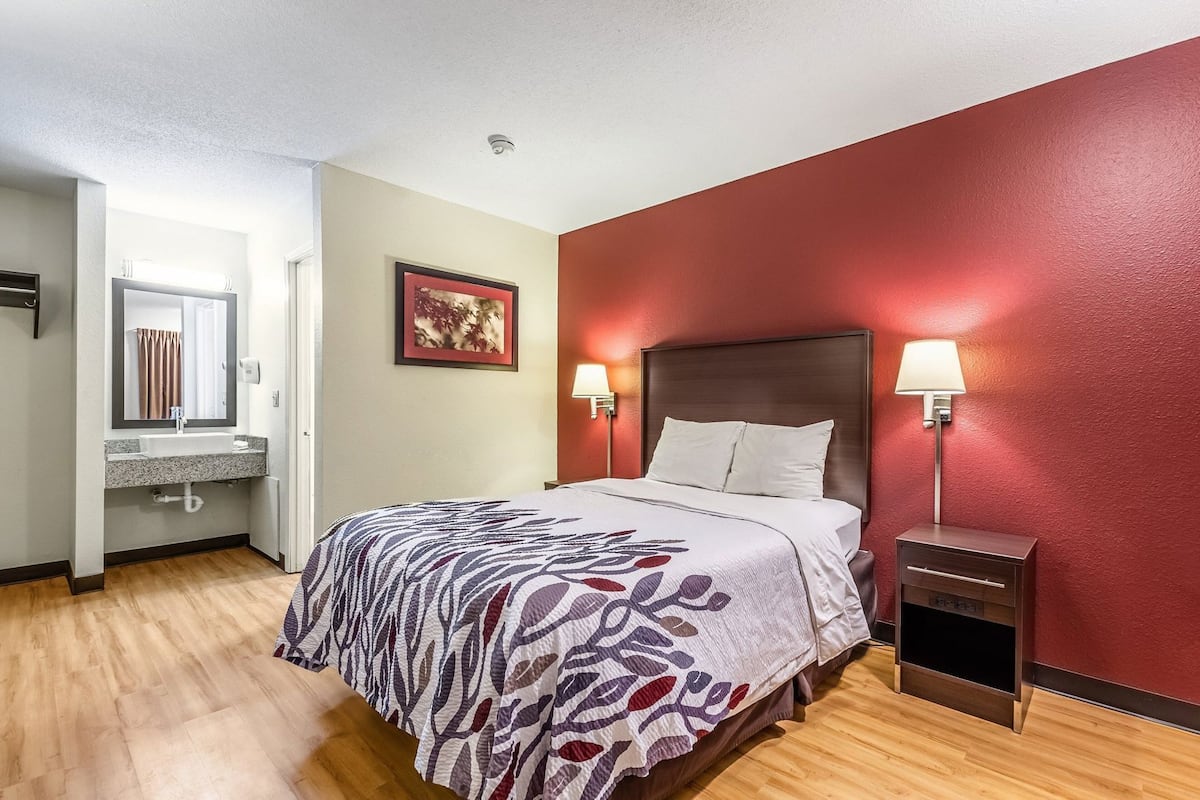 A hotel room with a bed, bedside lamps, a nightstand, a bathroom with a sink and mirror, and a red accent wall.