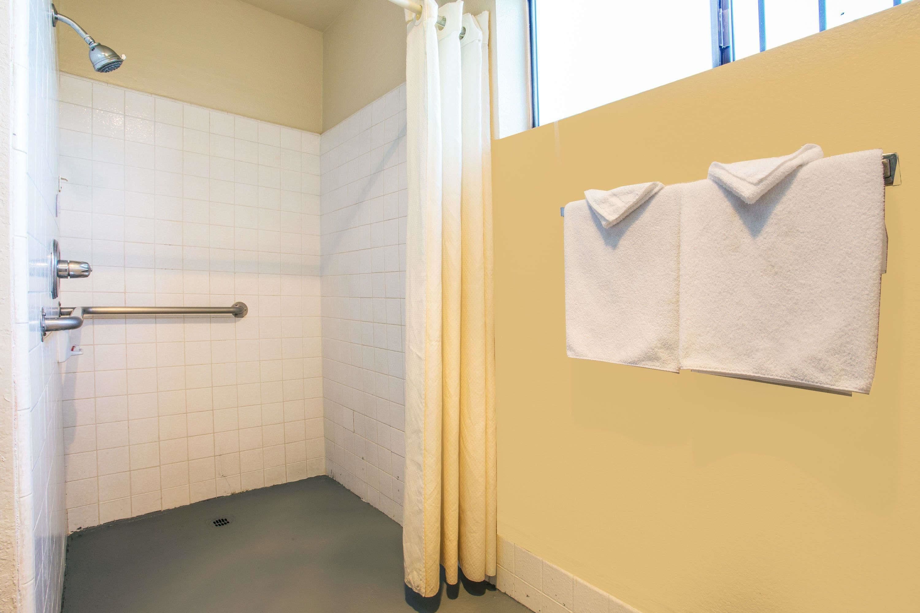 Ein kleiner, abgeschlossener Duschbereich mit weiß gefliester Wand, einer Duschbrause, einem Handtuchhalter mit zwei Handtüchern und einem Fenster mit Jalousien.