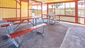 Un área de picnic techada con bancos y mesas, piso de concreto y un edificio amarillo y rojo al fondo.