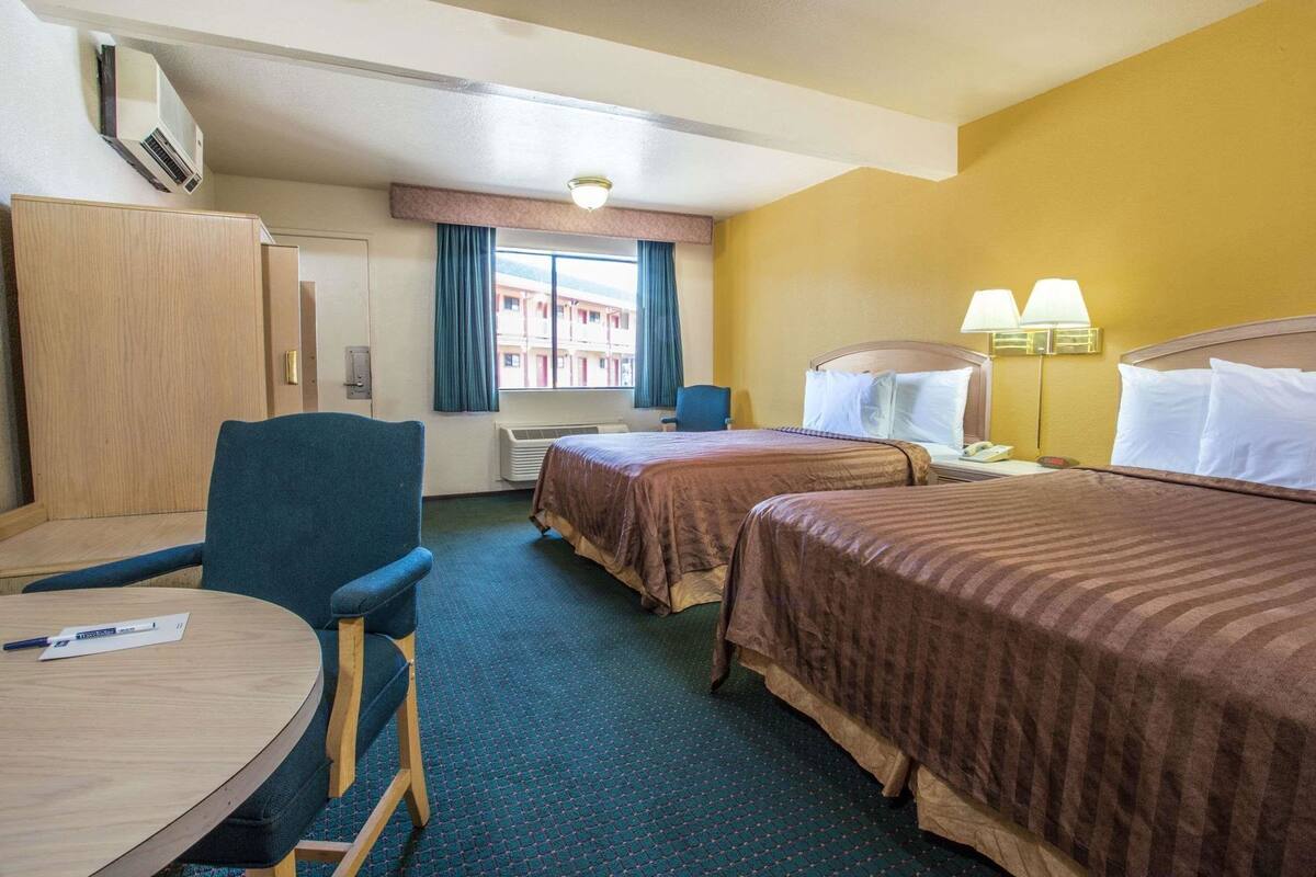 Une chambre d’hôtel avec deux lits, un bureau, une chaise, une lampe de bureau et une fenêtre avec des rideaux.