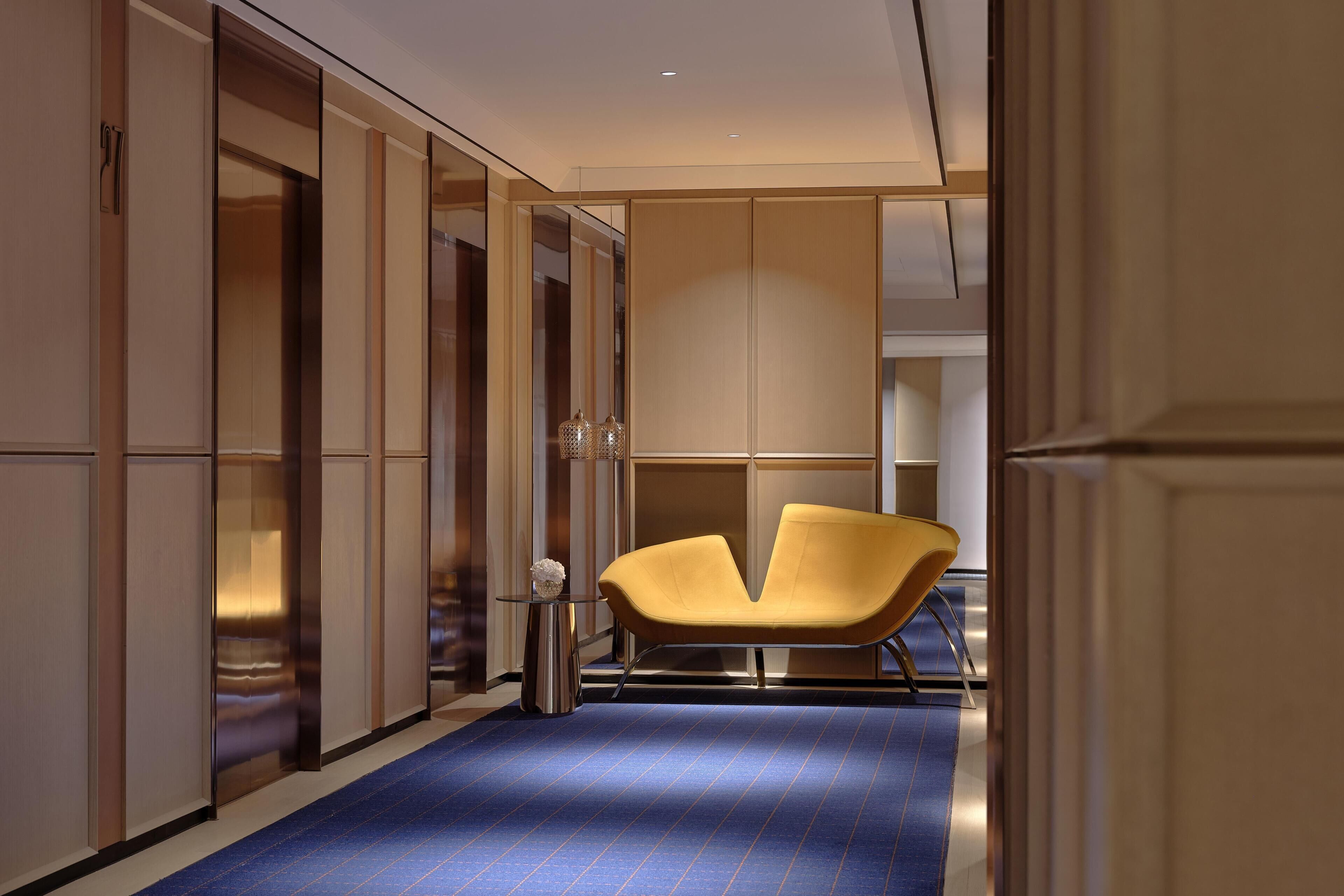 A modern hotel corridor with a blue carpet, a yellow chair, and a glass partition.