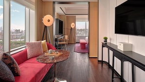 A modern hotel room with a large window, a sofa, a small table, a TV, and a desk.