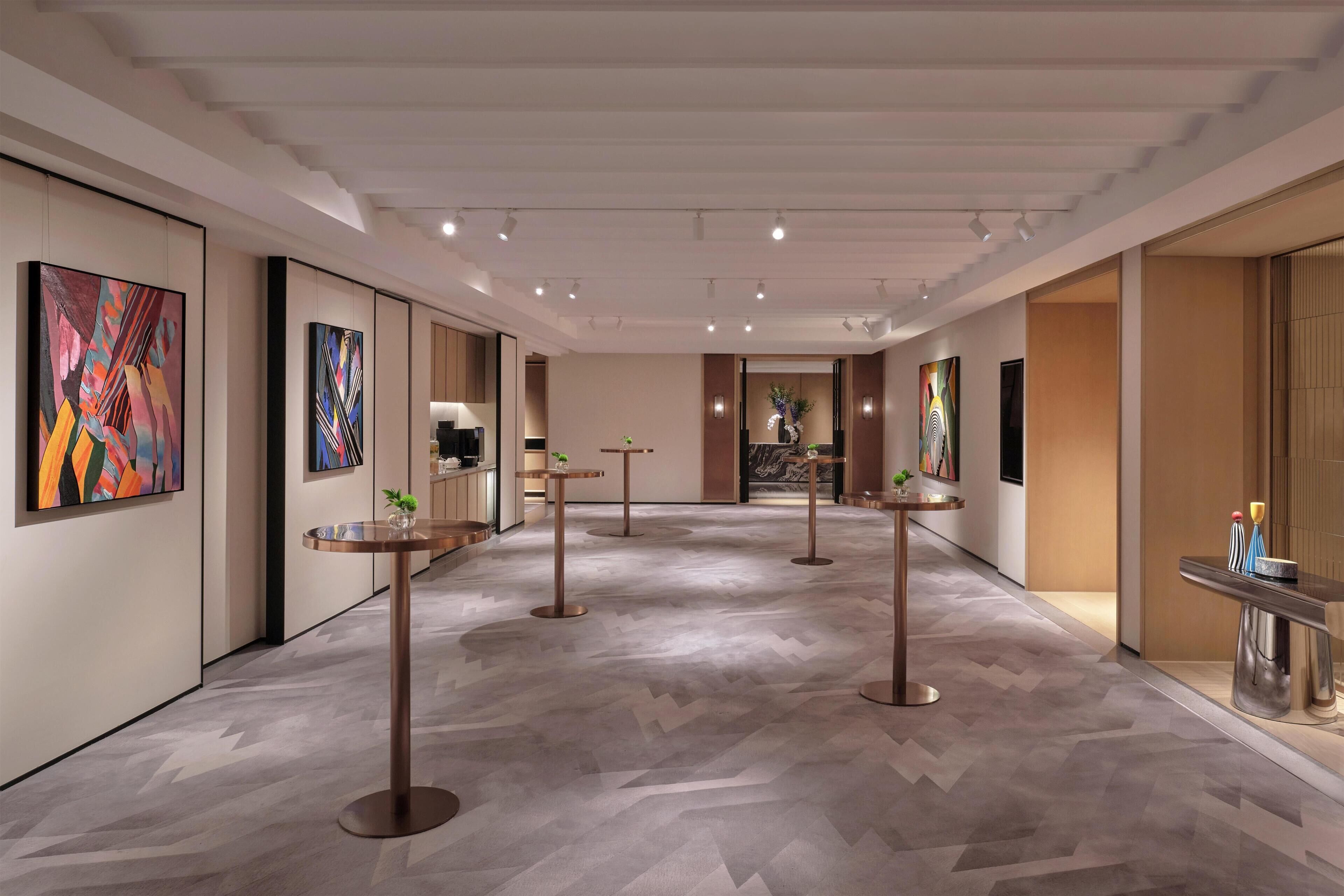 A modern hotel corridor with art on the walls, round tables, and a herringbone-patterned floor.