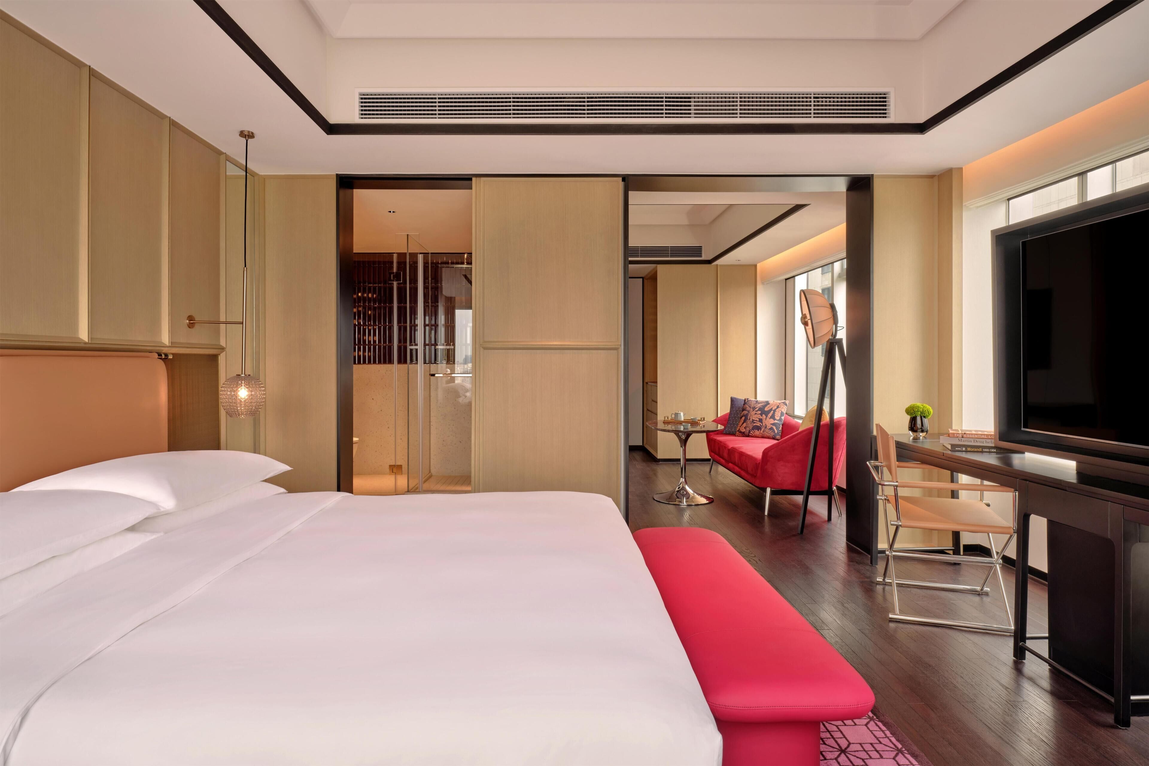 A hotel room with a large bed, a desk with a TV, a red sofa, and a dining area.