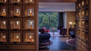 Un moderno vestíbulo de hotel con un amplio expositor de madera lleno de botellas de vidrio, una zona de estar con una mesa redonda, y vistas al exterior a través de grandes ventanales.