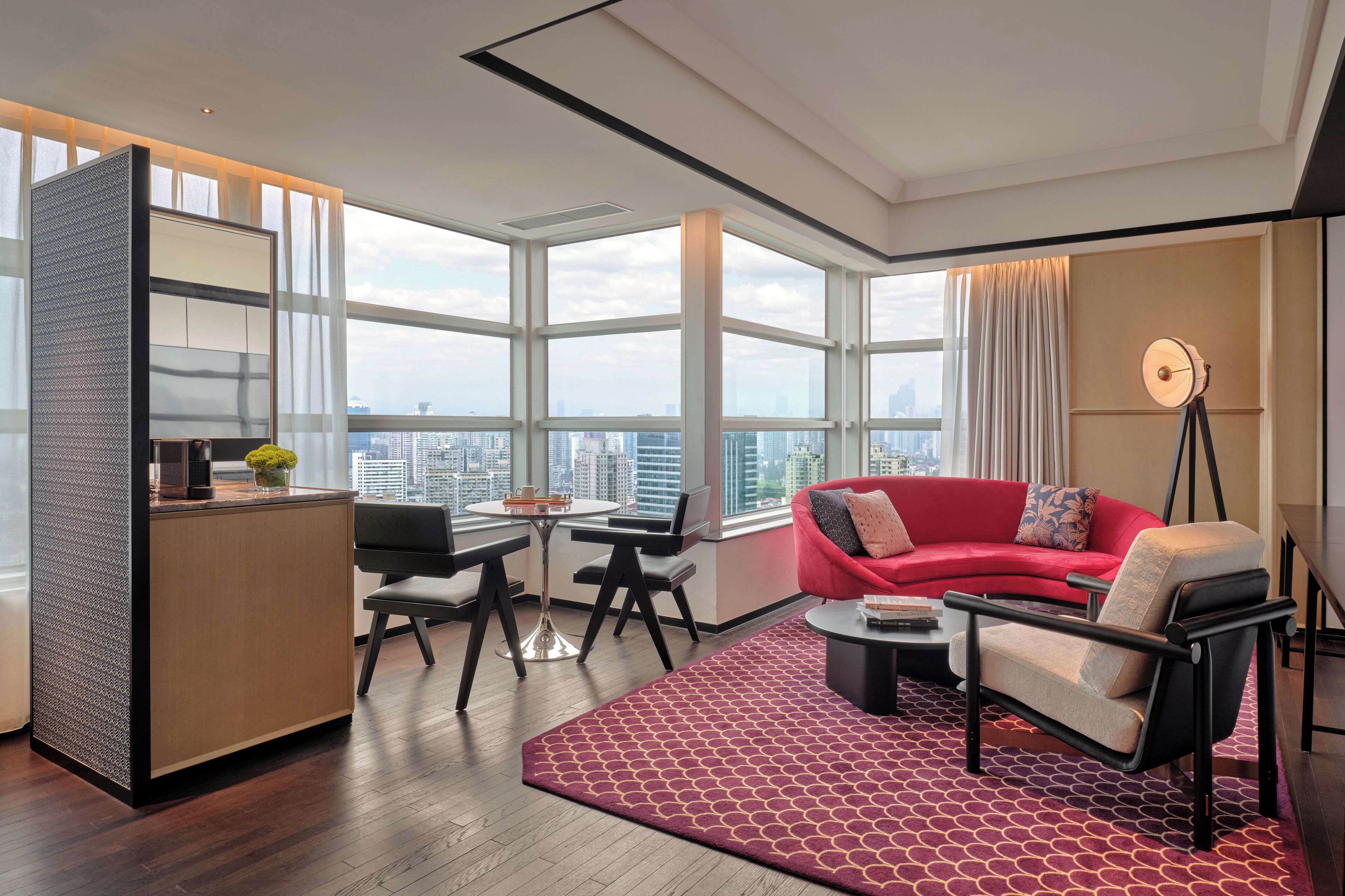 A modern living room with a red sofa, a coffee table, a desk with a chair, and a large window offering a city view.