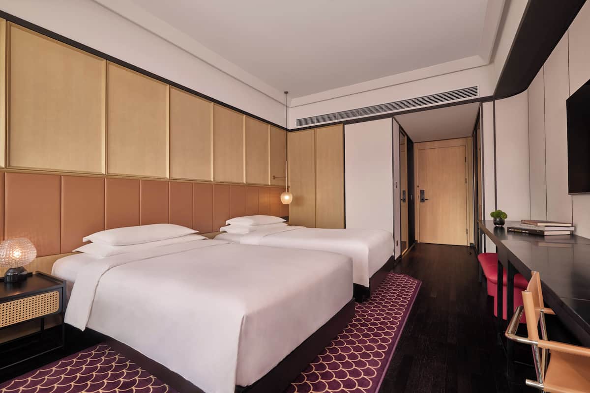 A hotel room with two beds, a desk, a chair, a TV, and a plant.