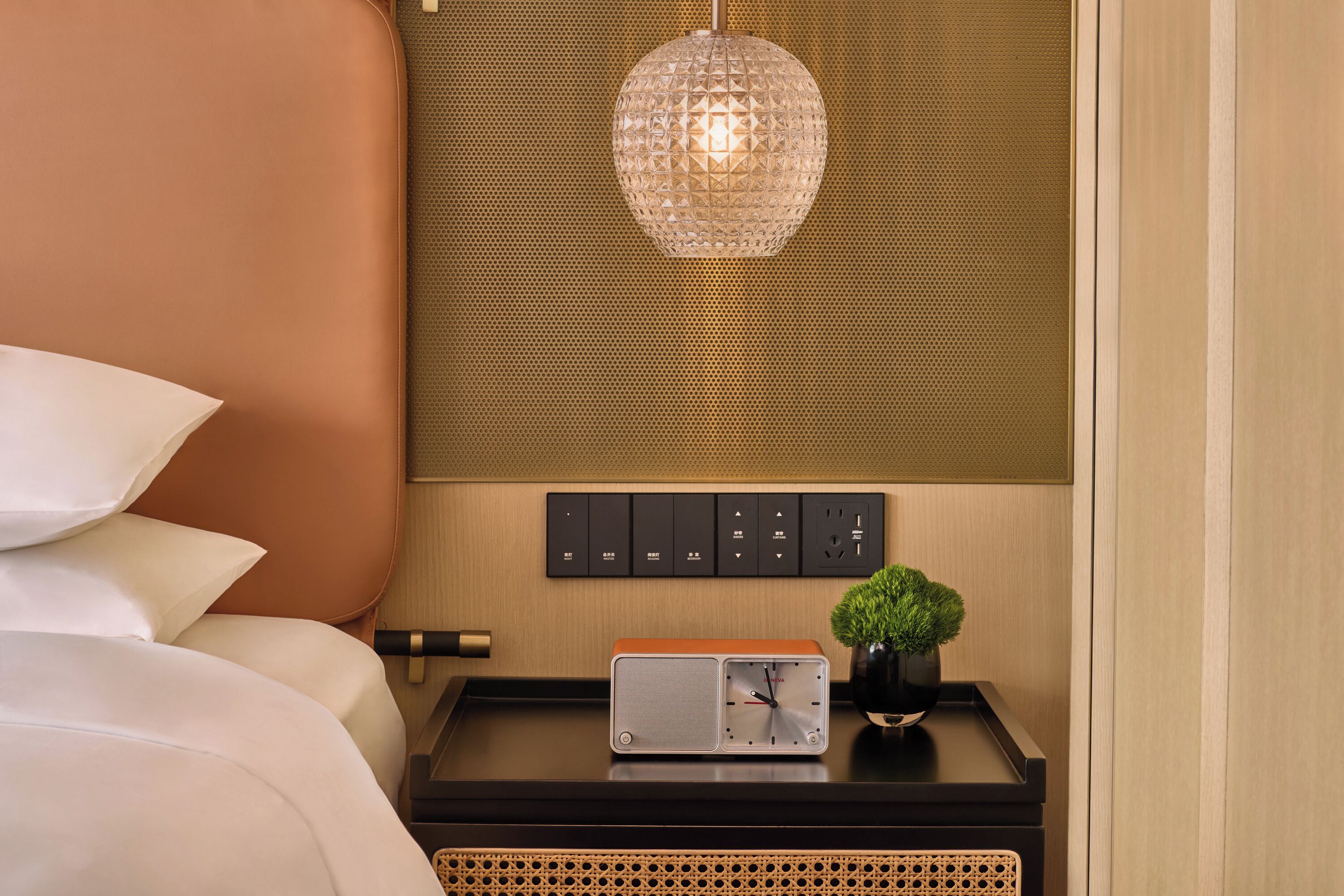 Quarto de hotel bem arrumado com uma cama, uma mesa de cabeceira com um relógio, uma pequena planta e um painel de controle fixado na parede.