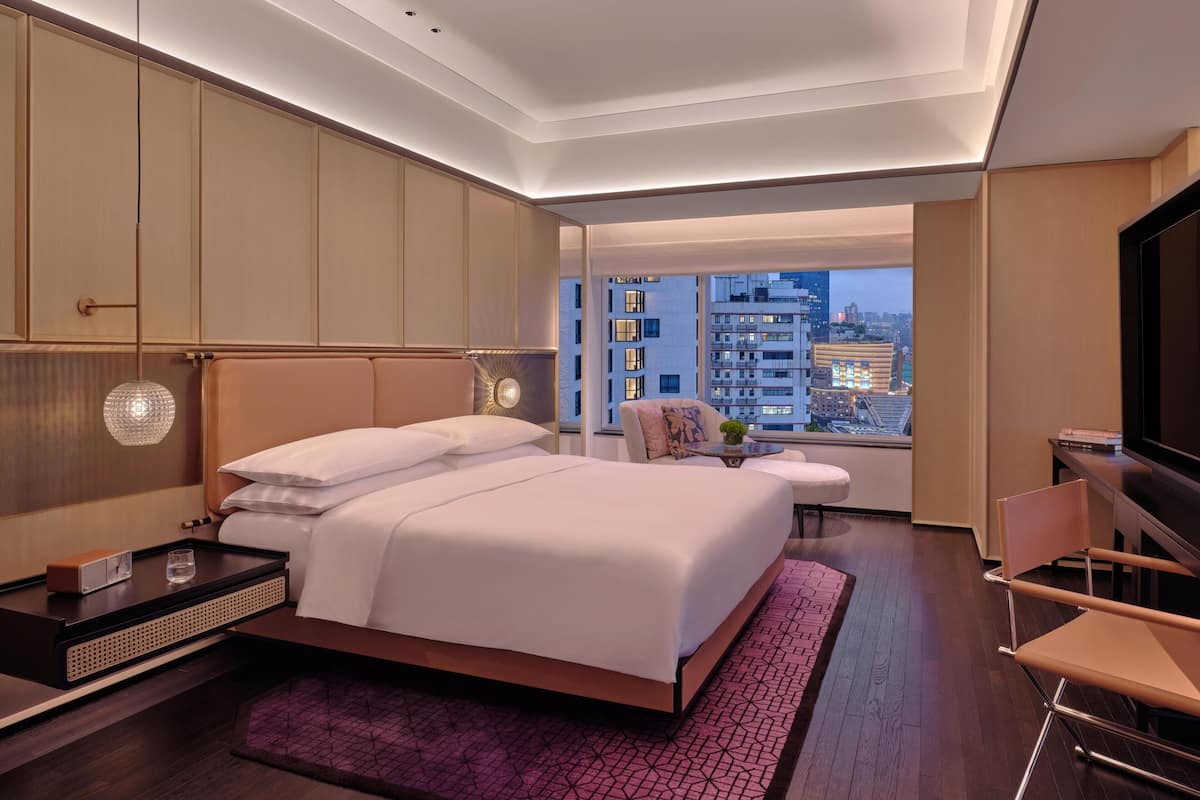 A modern hotel room with a large bed, a TV, a desk, and a view of the cityscape.