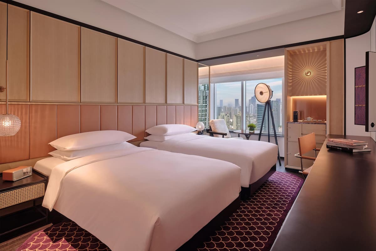 A hotel room with two beds, a desk, a clock, and a city view.