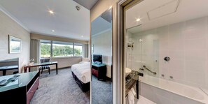 A hotel room with a bed, desk, chair, and a glass-enclosed shower area.