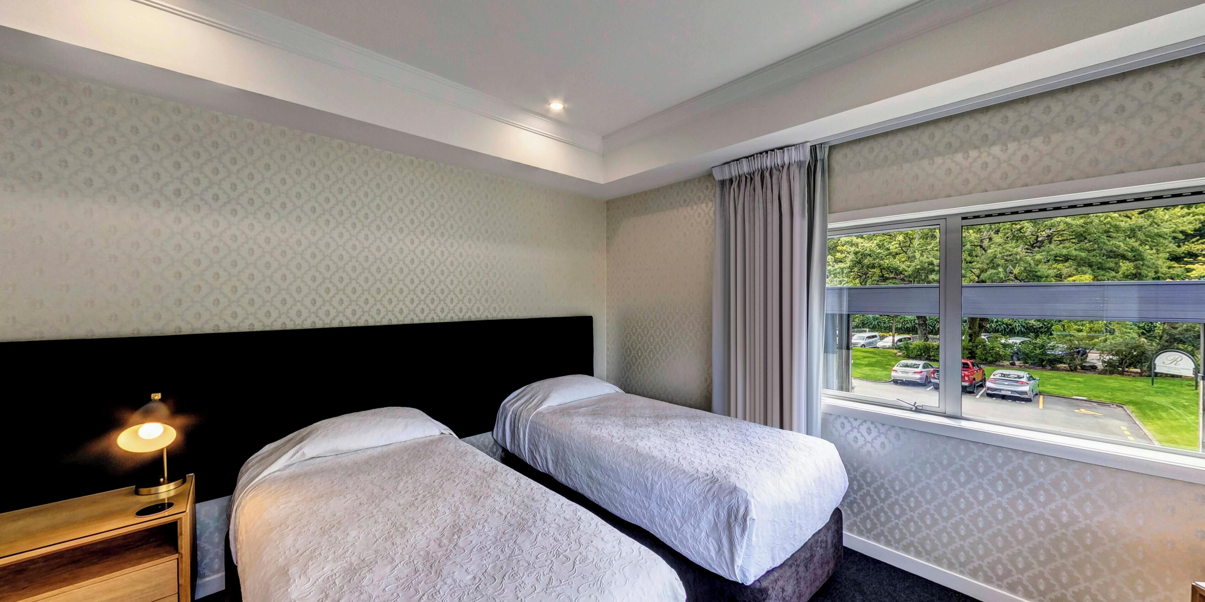 A hotel room with two beds, a nightstand with a lamp, and a window with a view of a parking lot.