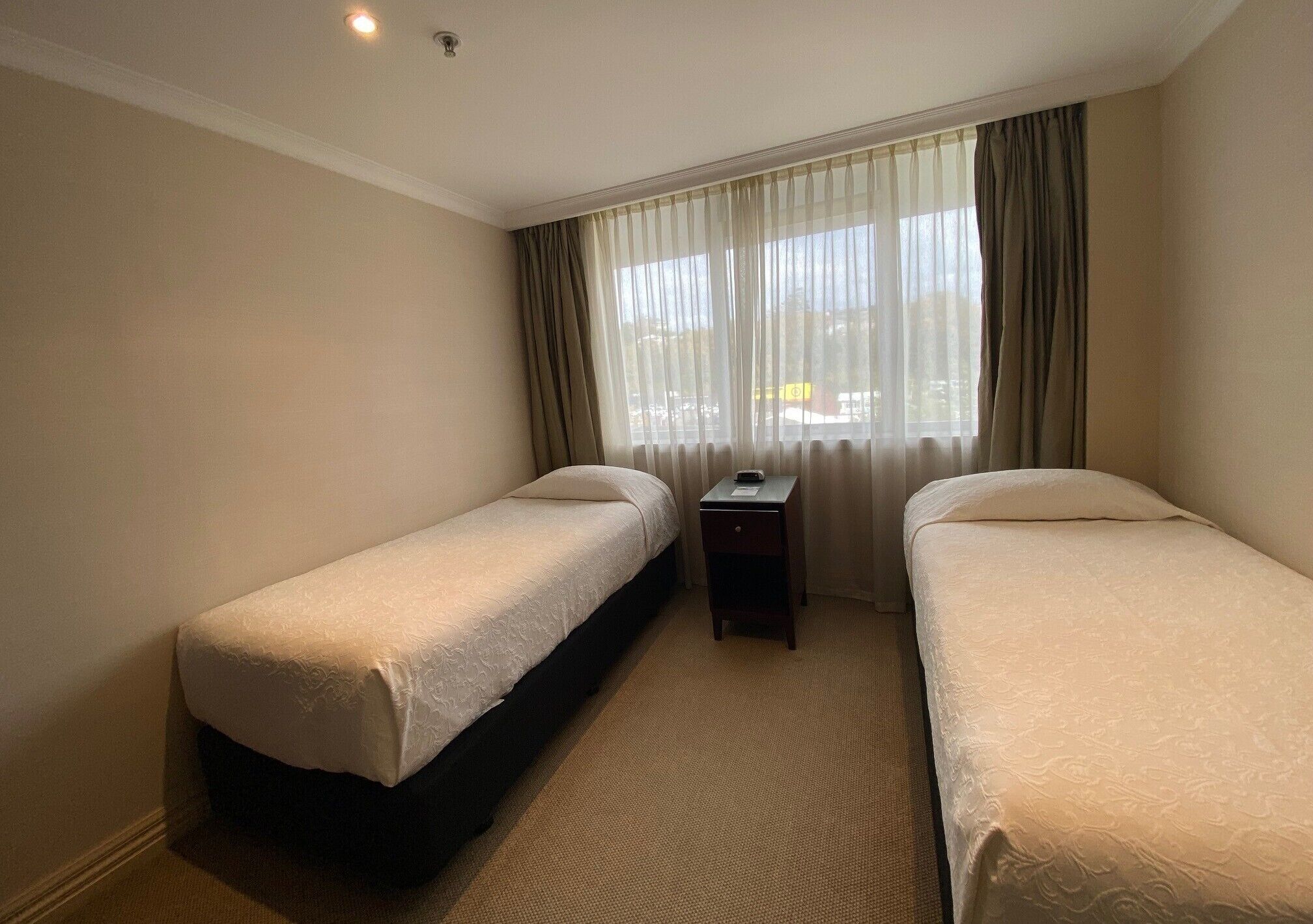 A hotel room with two single beds, a small bedside table, and a window with curtains.