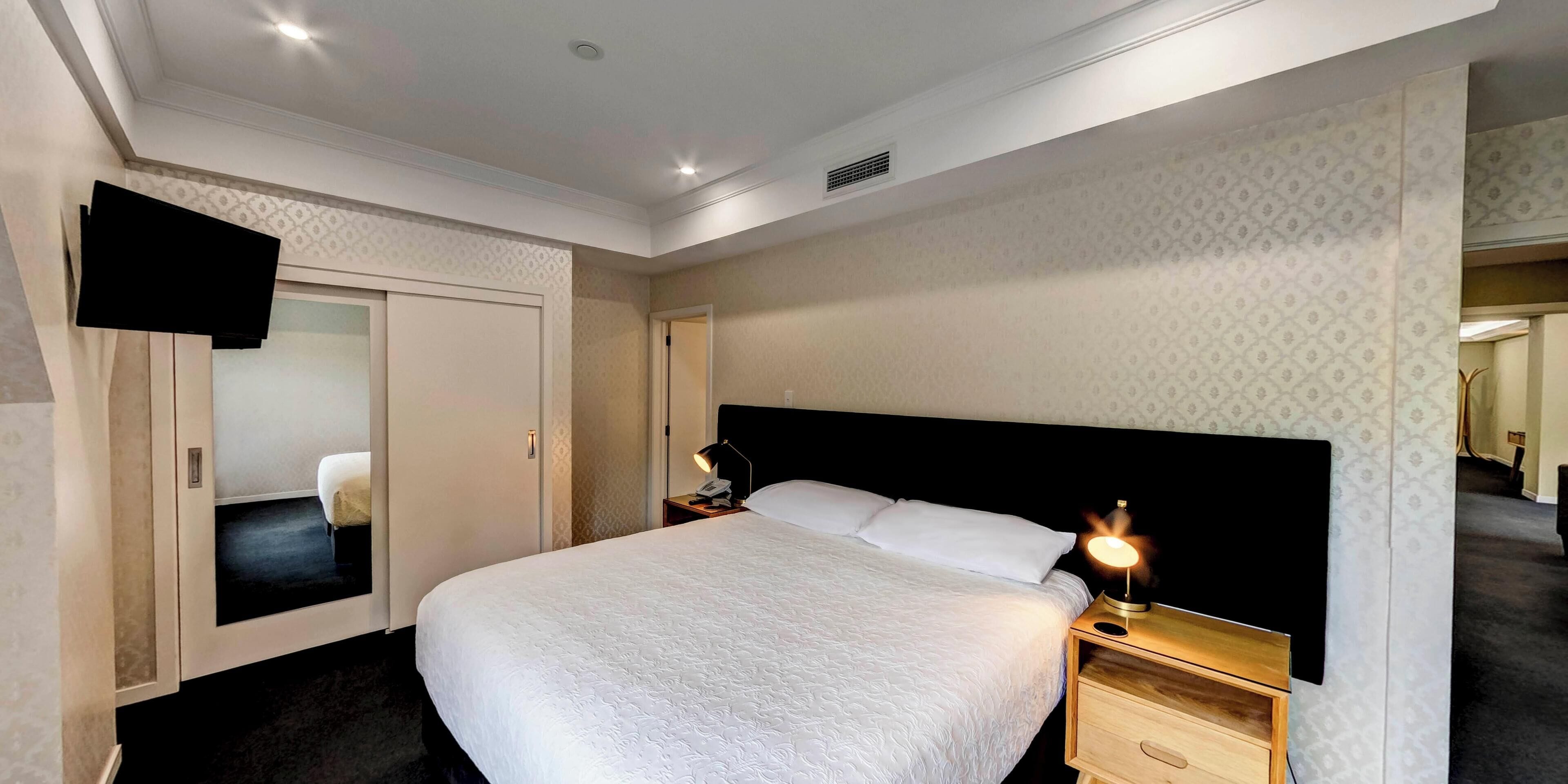 A hotel room with a large bed, a bedside table with a lamp, a wardrobe, and a television mounted on the wall.