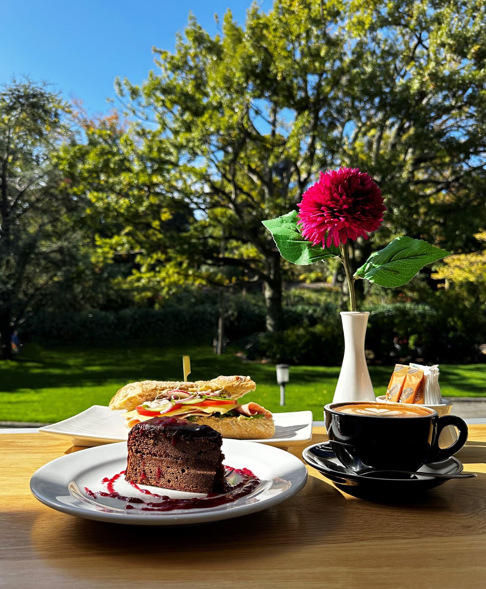 A table with a dessert, a sandwich, and a cup of coffee, set outdoors with a garden view.