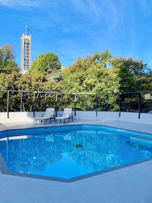 A swimming pool with lounge chairs, surrounded by trees and a tall building in the background.