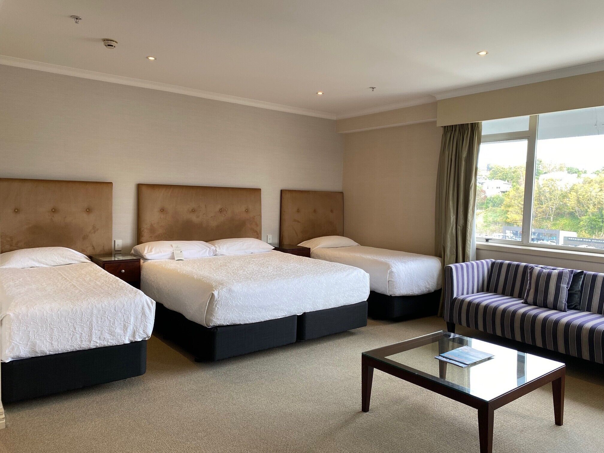 A hotel room with two beds, a sofa, a glass coffee table, and a large window with a view.