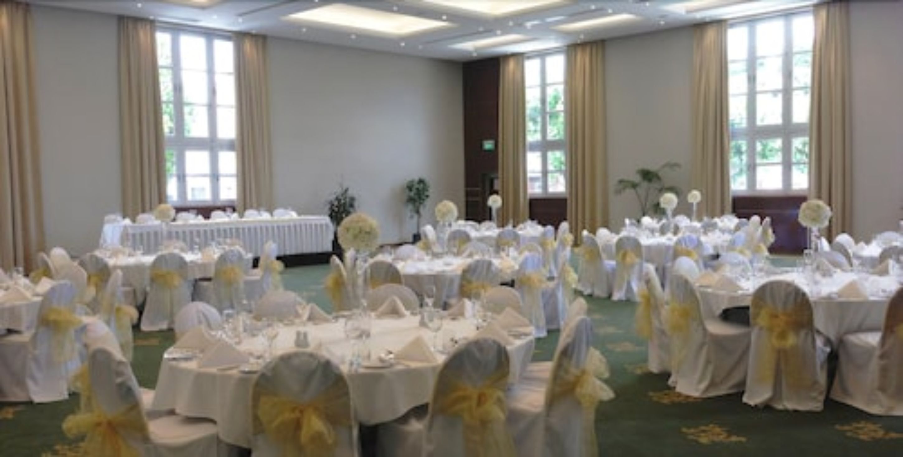 Un salón de banquetes con mesas redondas cubiertas con manteles blancos y sillas adornadas con cintas amarillas, dispuesto para un evento formal.