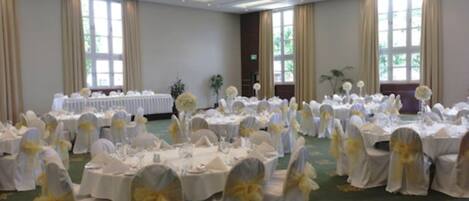 Un salón de banquetes con mesas redondas cubiertas con manteles blancos y sillas adornadas con cintas amarillas, dispuesto para un evento formal.