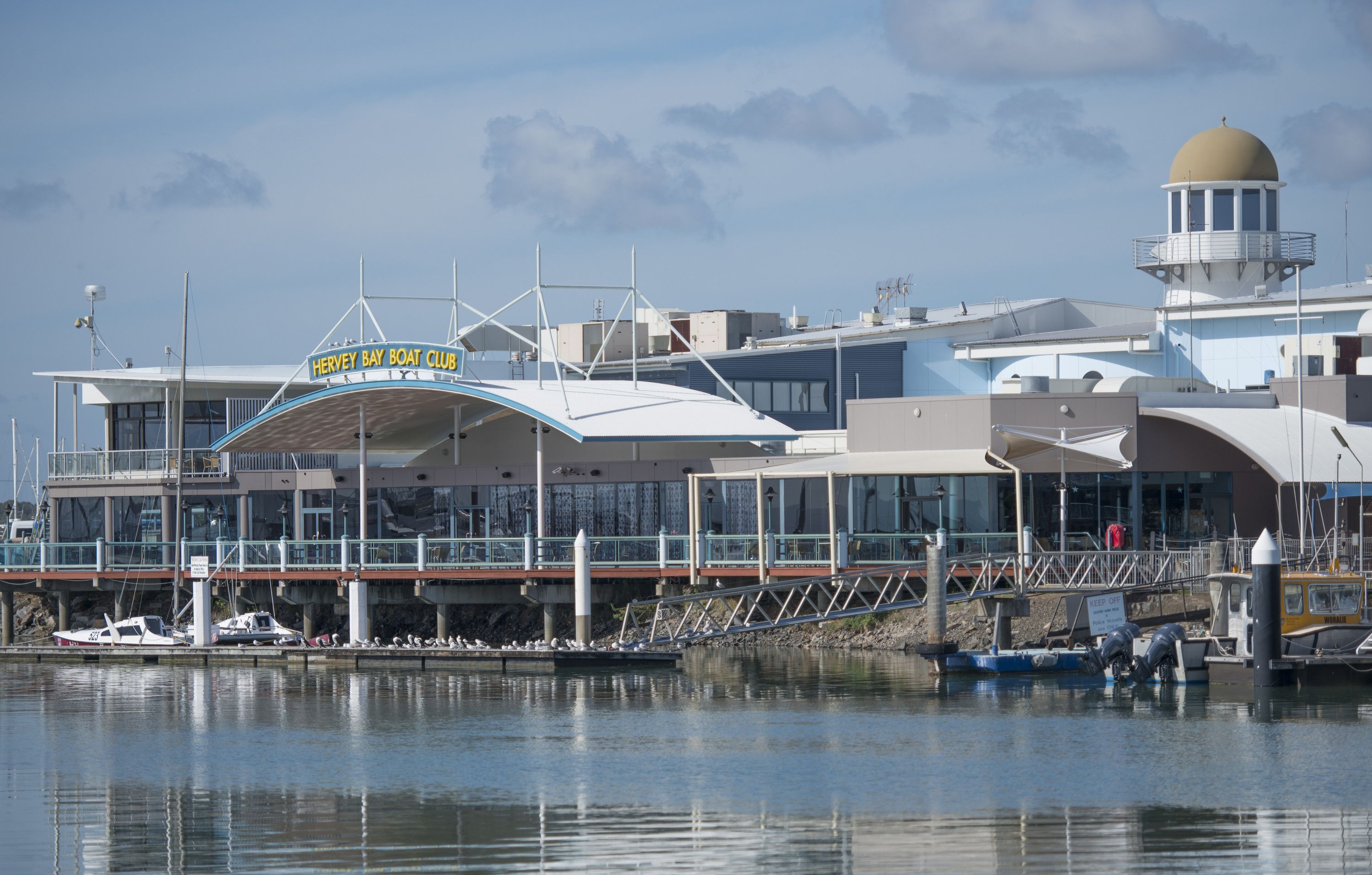Un port de plaisance avec des bateaux amarrés, un hangar à bateaux et un bâtiment surmonté d’un dôme et portant une enseigne indiquant « HERVEY BAY BOAT CLUB ».