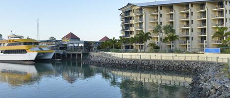 Un puerto deportivo con un yate grande amarrado, un hotel de varios pisos con balcones y una costa rocosa.