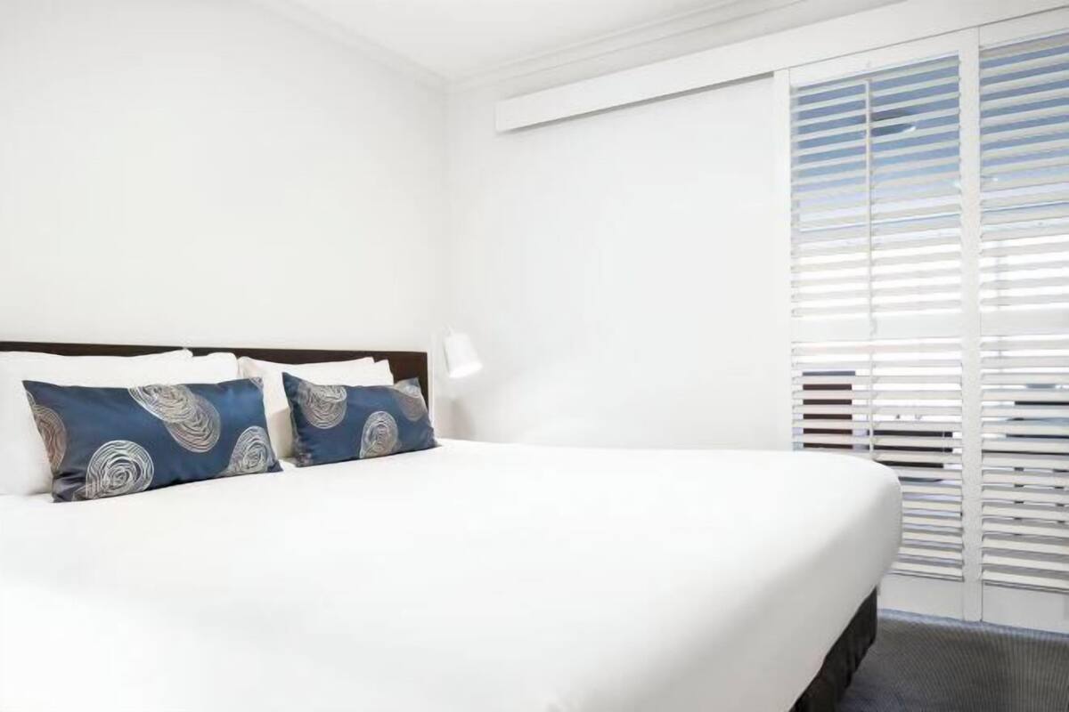 Une chambre d’hôtel avec un lit, deux oreillers, une table de chevet et une fenêtre avec des stores.