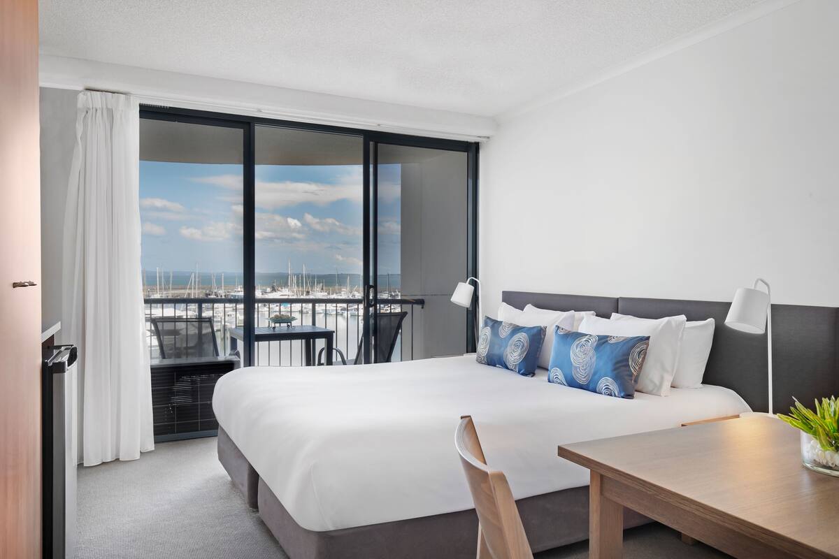 Une chambre d’hôtel avec un grand lit, un bureau, une chaise et un balcon donnant sur le port de plaisance.