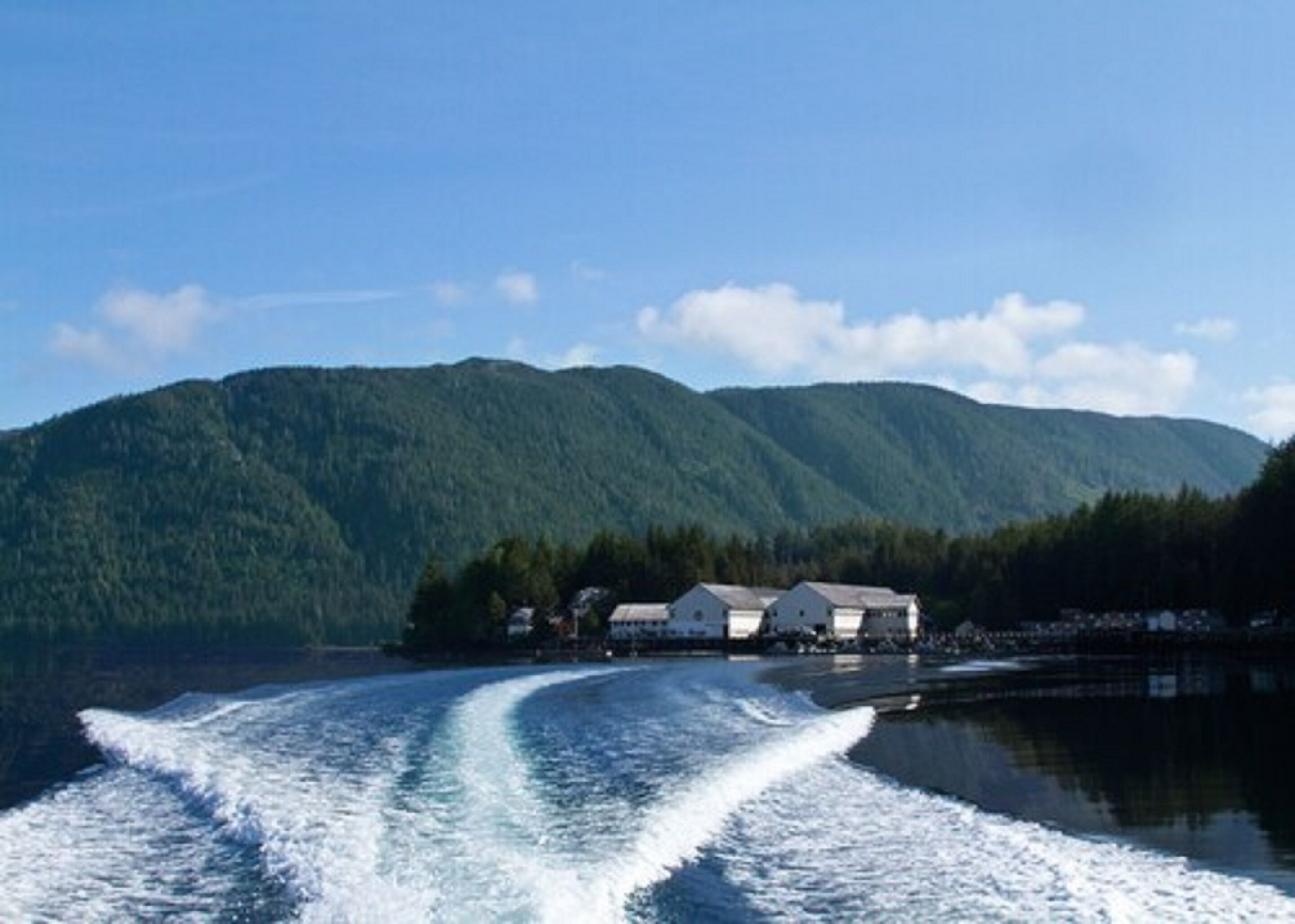 Ein Boot fährt auf einem See, im Hintergrund Berge und eine Reihe von Gebäuden.