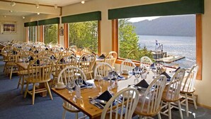 Un comedor con mesas y sillas de madera, ventanas amplias con vista a un lago y montañas, y un pequeño espacio de asientos al aire libre visible a través de las ventanas.