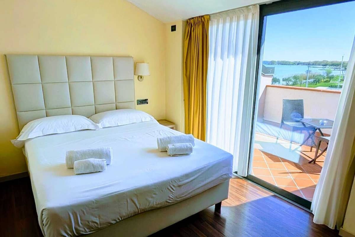 A hotel room with a large bed, a view of the outdoors through a sliding glass door, and a balcony with outdoor furniture.
