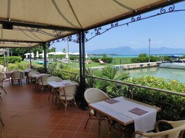 A covered outdoor seating area with tables and chairs, overlooking a body of water and mountains.