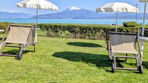 Un espace extérieur verdoyant, agrémenté de parasols blancs et de chaises longues, offrant une vue sur un plan d’eau et des montagnes.