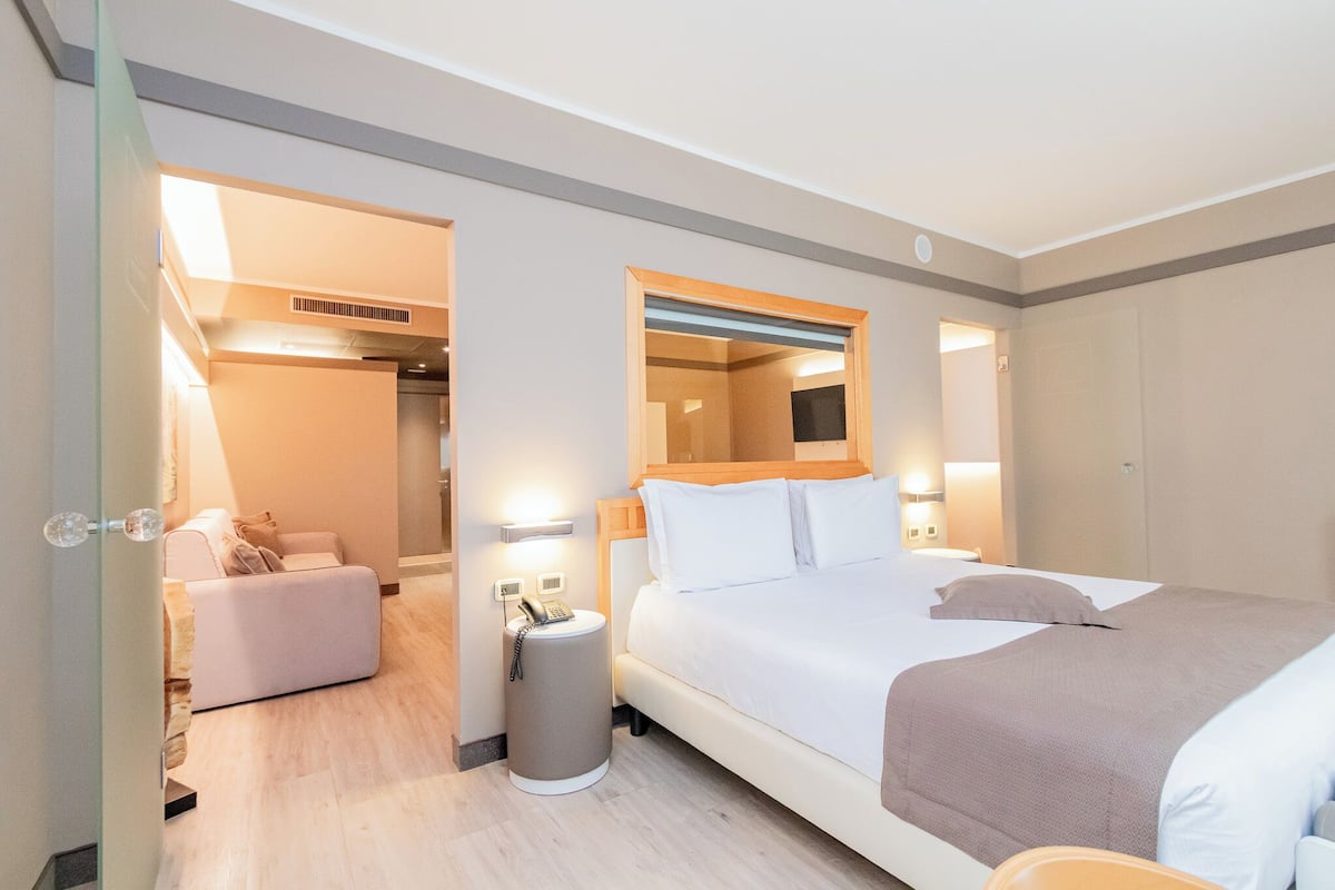 A hotel room with a bed, bedside table, lamp, and a small seating area with a chair and a table.