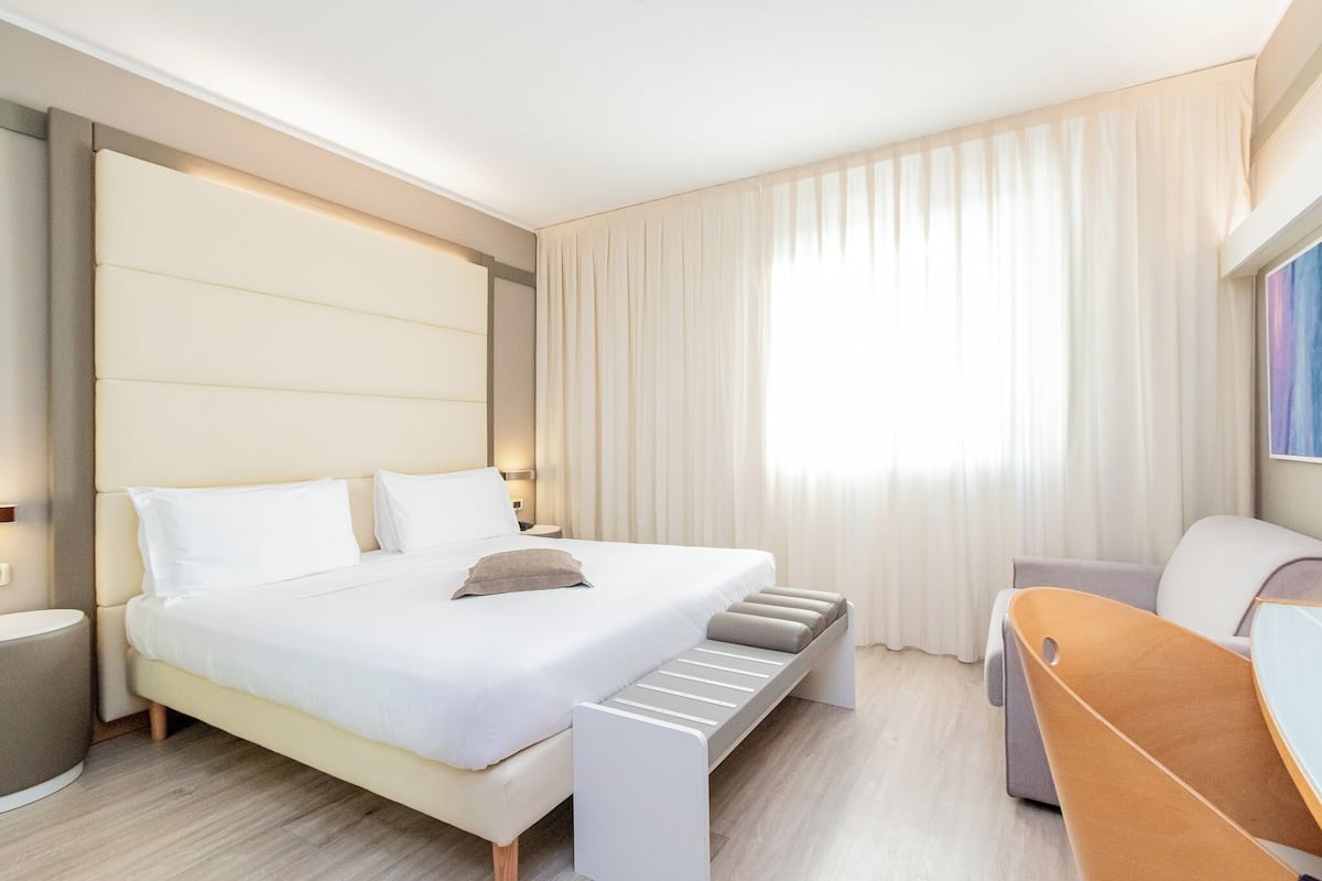 A modern hotel room with a large bed, a bedside table, a chair, and a window with curtains.
