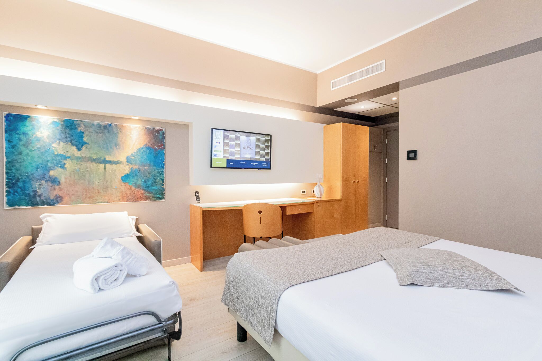 A hotel room with two beds, a desk, a large abstract painting, and a digital clock on the wall.