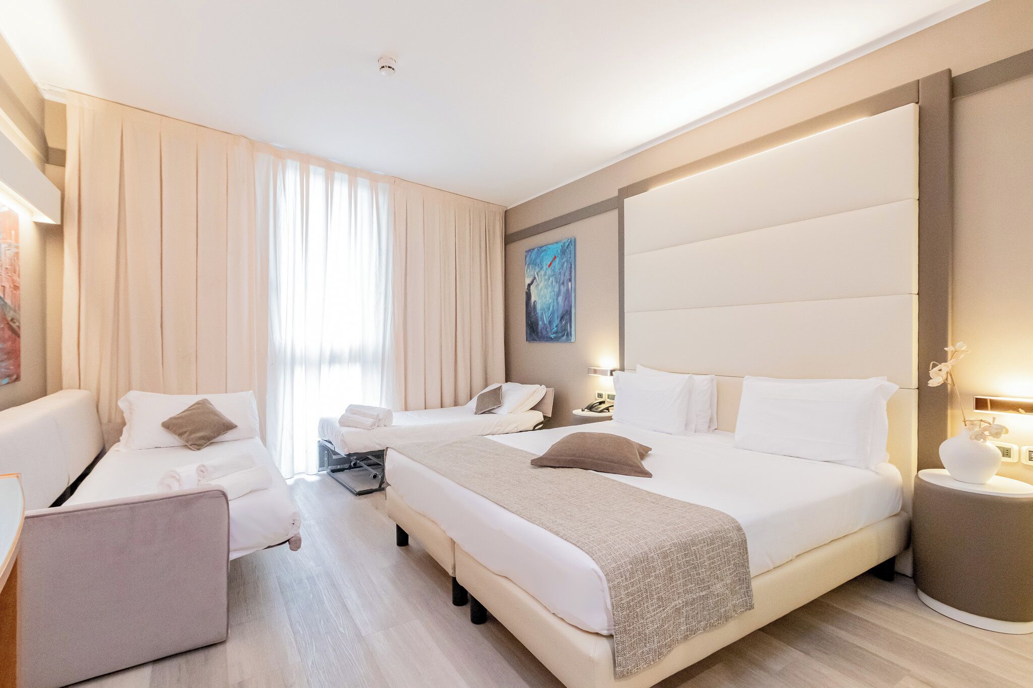 A modern hotel room with a large bed, two bedside tables, a seating area with a chair, and a large window with curtains.