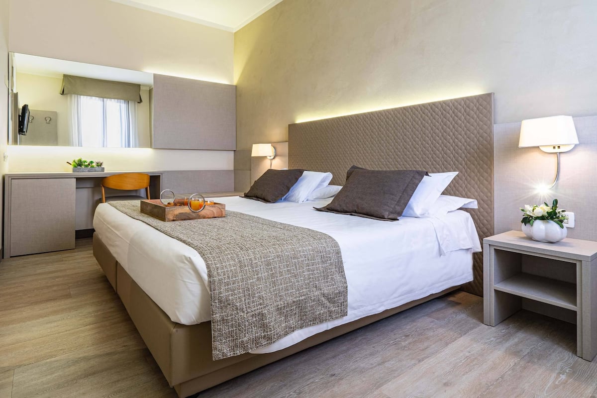 A modern hotel room with a large bed, bedside tables, a desk, and a mirror.