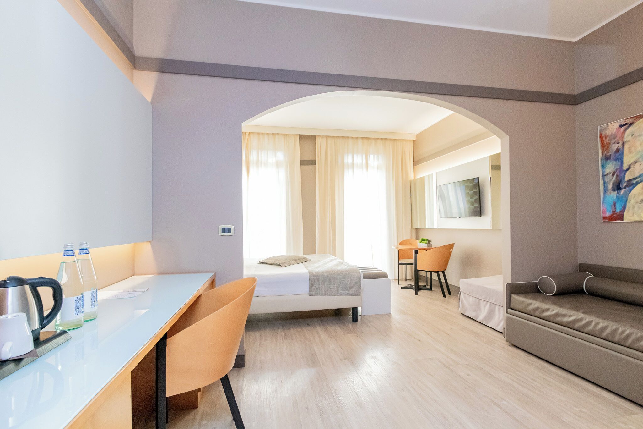 A modern hotel room with a bed, a desk, a chair, a sofa, and a large window with curtains.