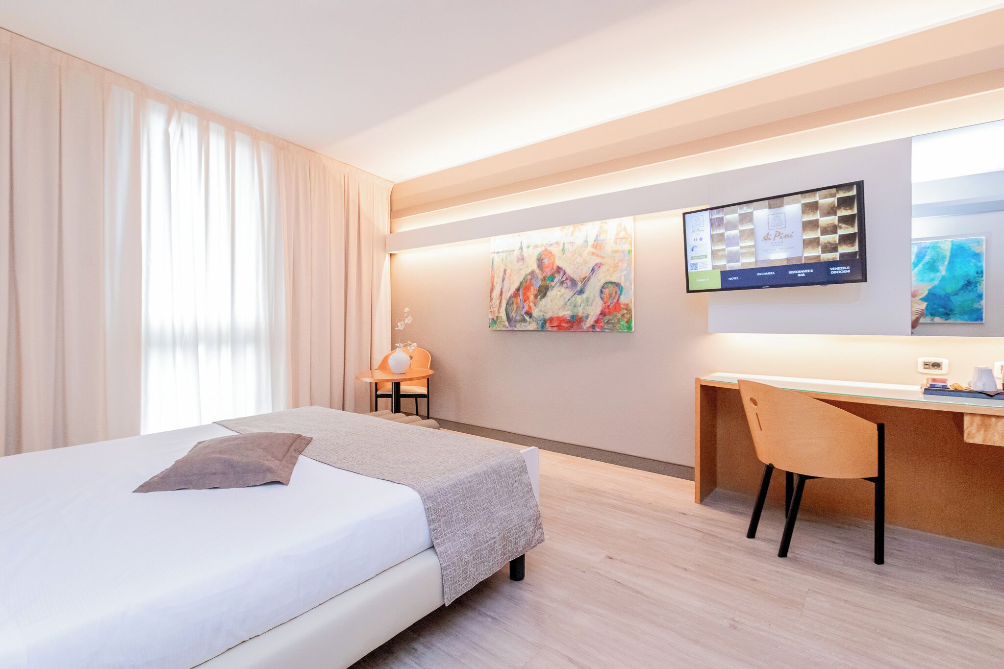 A hotel room with a bed, a desk, and a TV. There are curtains, a small table with a lamp, and artwork on the walls.
