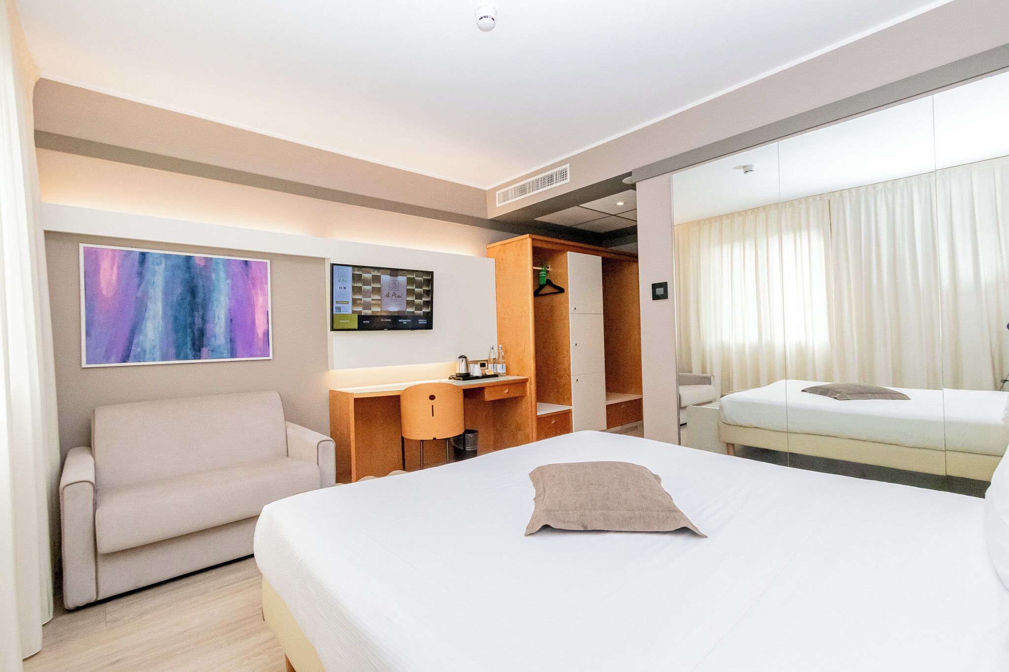 A modern hotel room with a bed, a desk, a sofa, and a wardrobe.