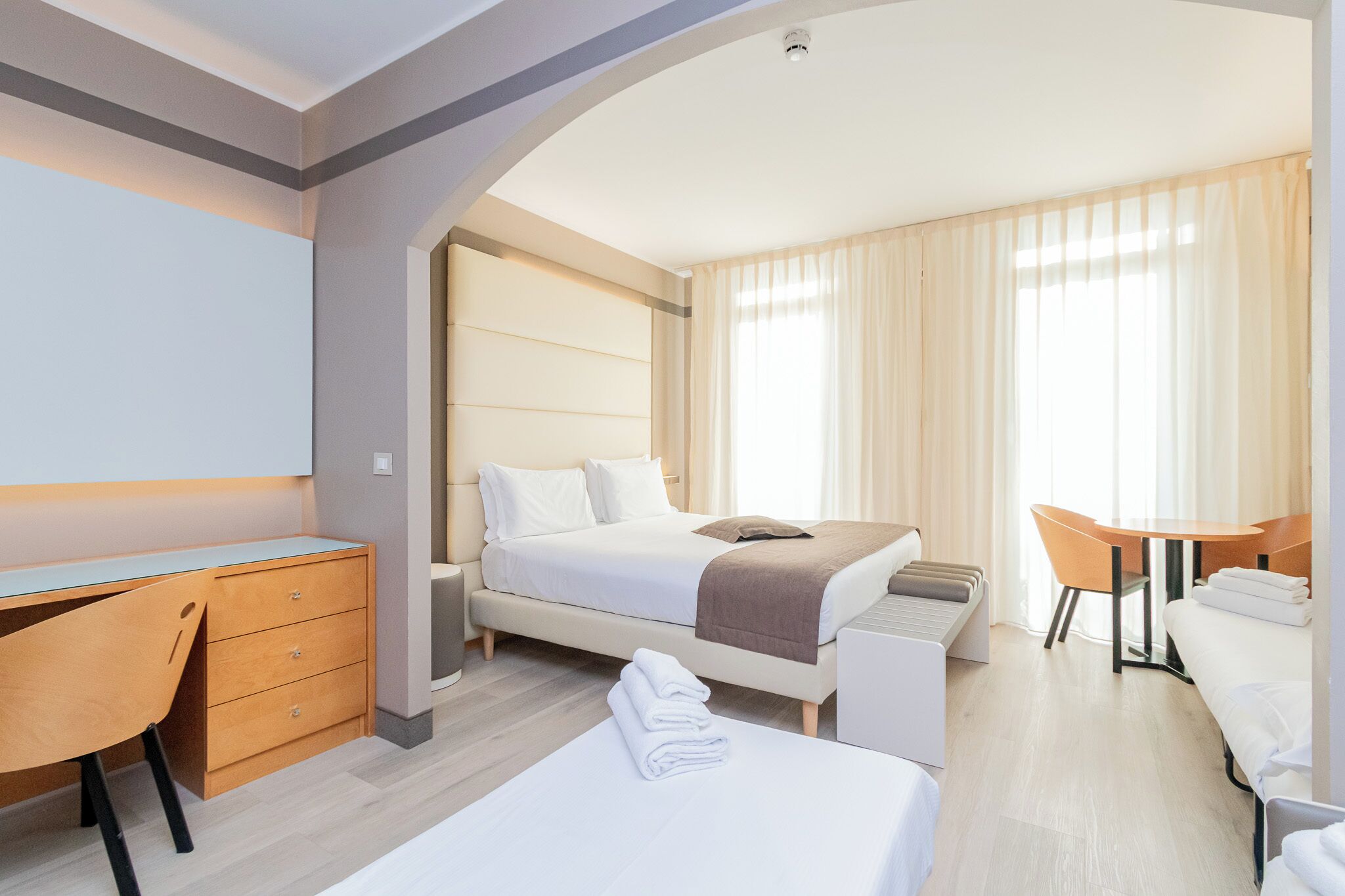 A hotel room with a bed, a desk, a chair, a small table, and a bench.