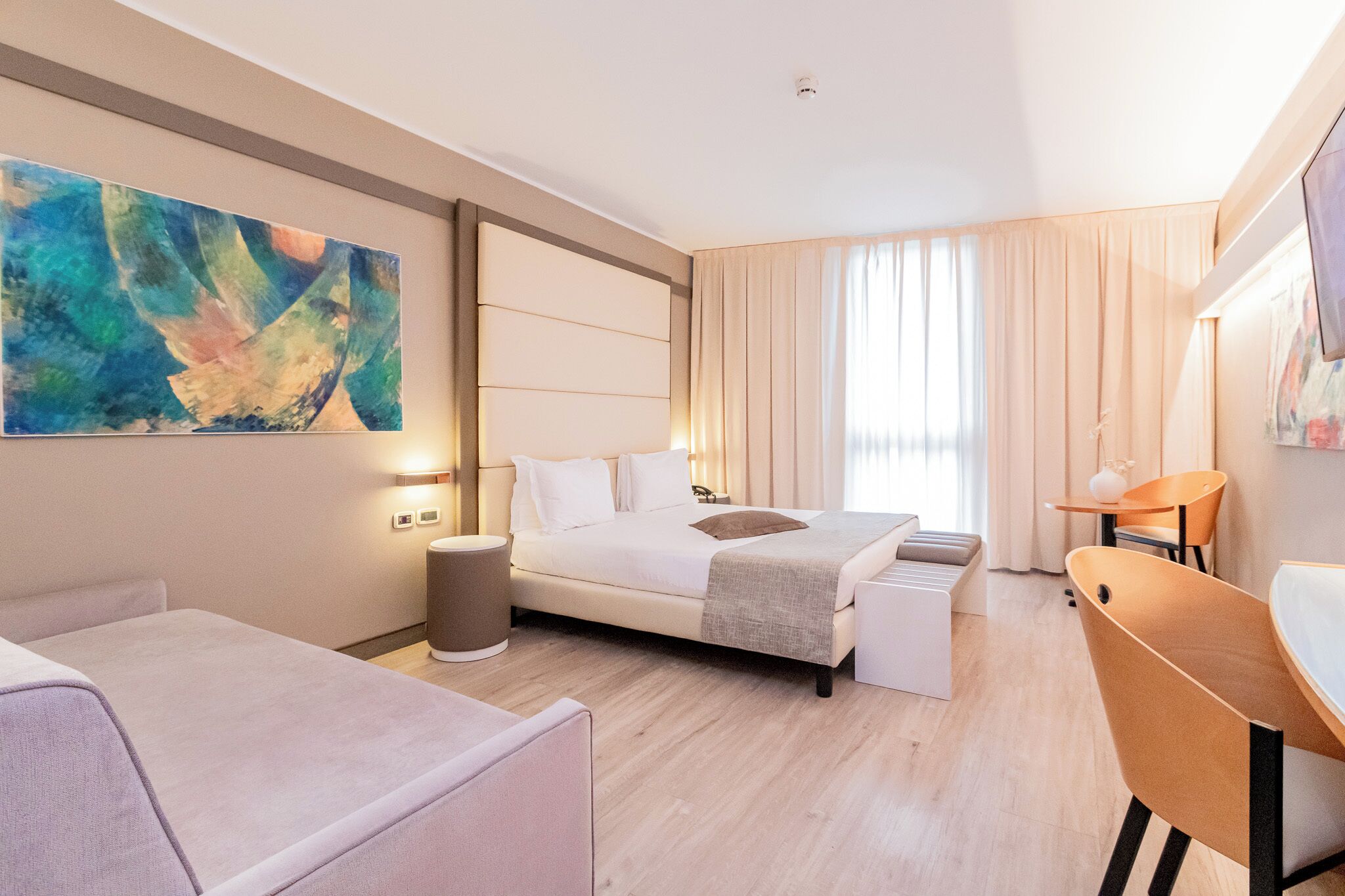 A modern hotel room with a large bed, a desk, and a chair. There is a painting on the wall and a window with curtains.