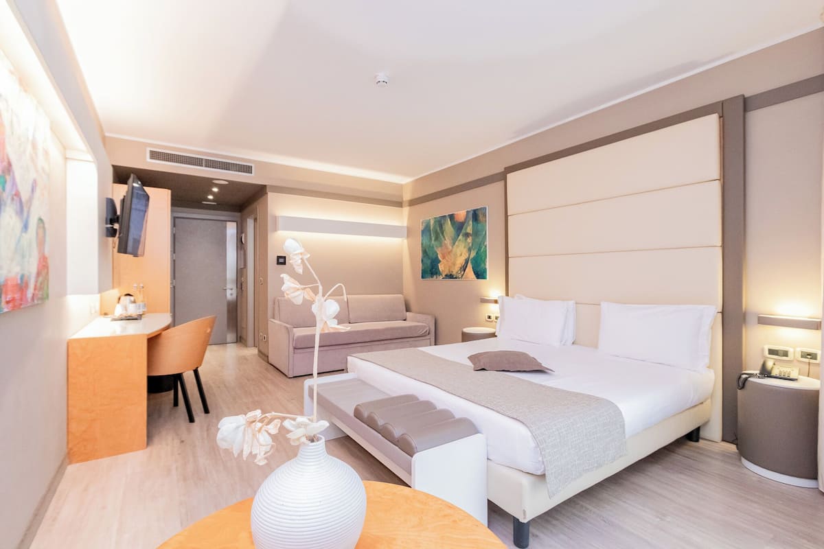 A modern hotel room with a large bed, a desk, a sofa, and a dining area.