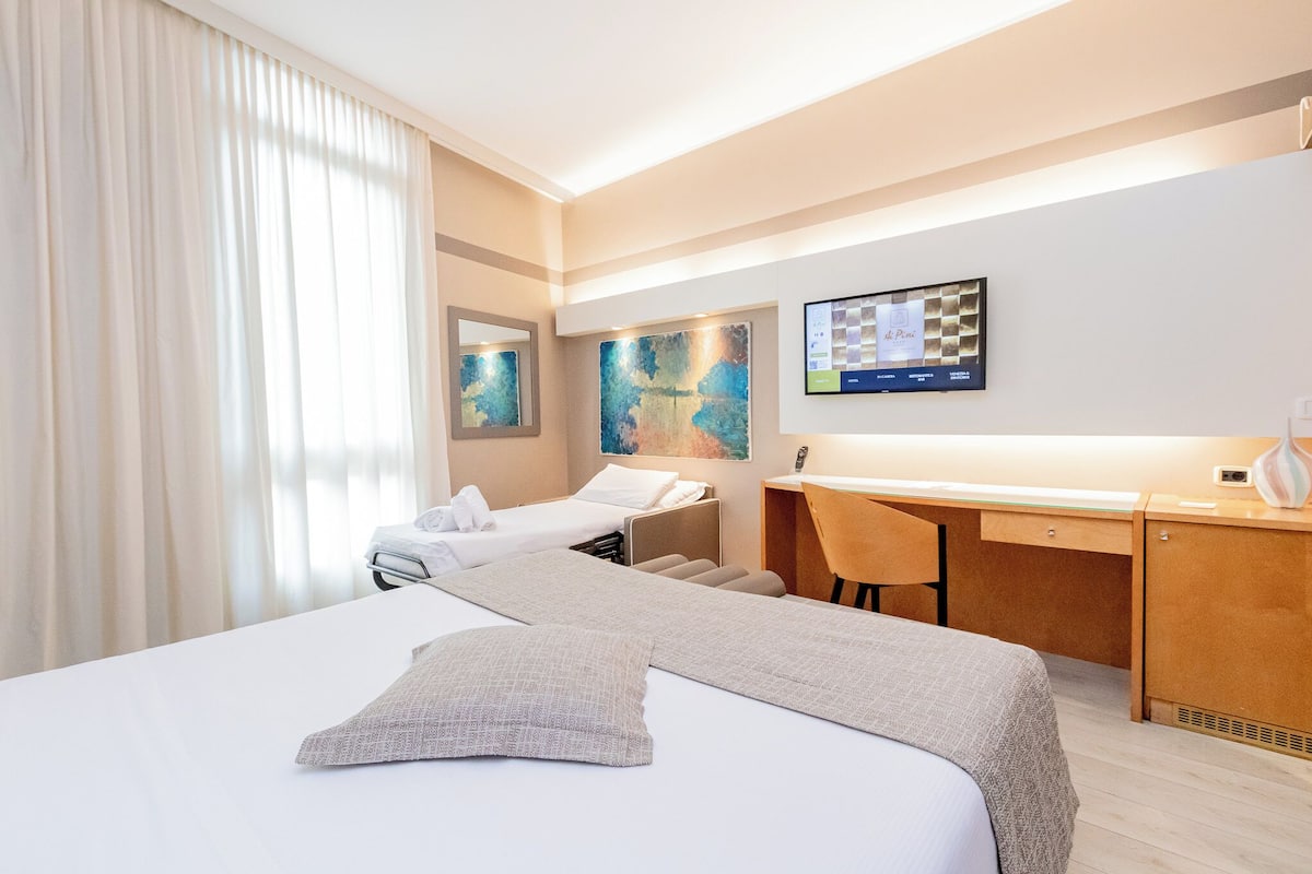 A hotel room with a large bed, a desk, and a chair. There is a television on the wall and artwork above the desk.